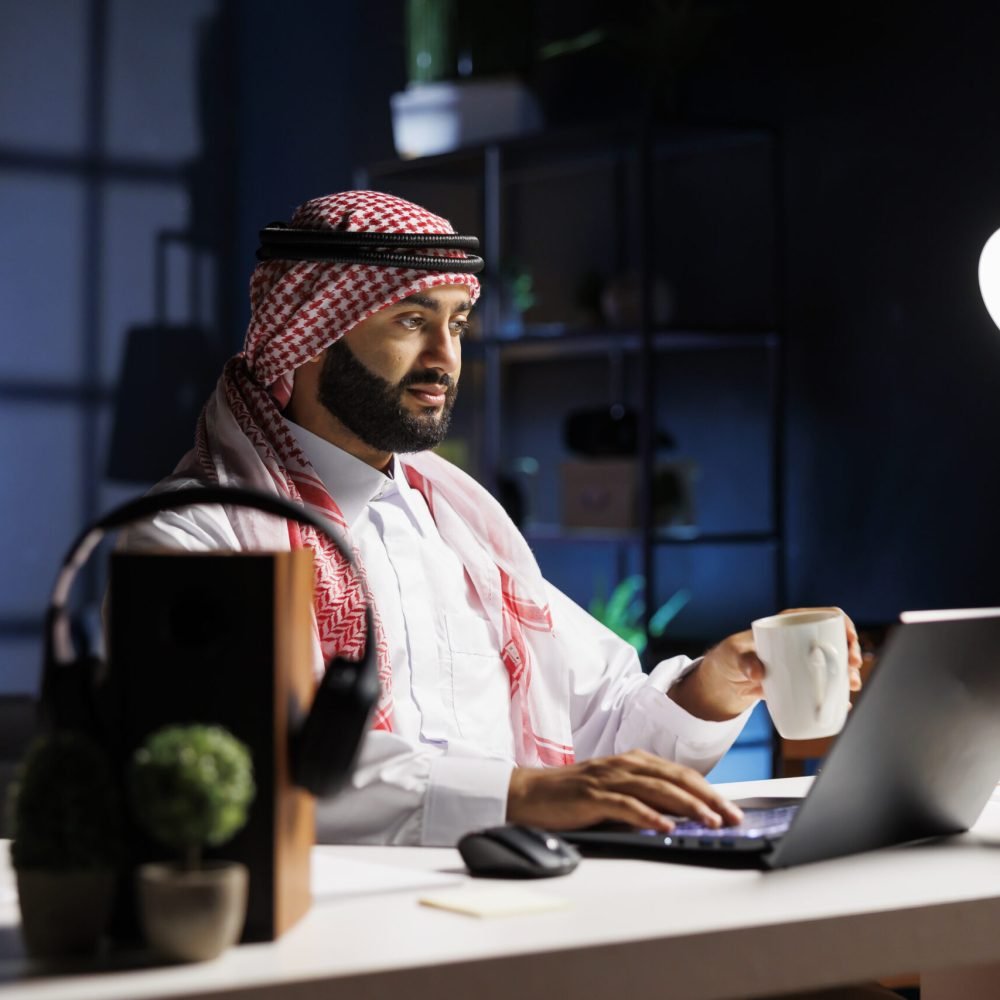 Side-view of a Muslim guy holding a cup of coffee and surfing the net on his digital laptop. Image showcasing a male Arab person engrossed in his personal computer while drinking tea.